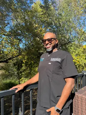 Dr. Macklin Lacy, a smiling man in a gray shirt with the words 'Dr. Macklin Lacy, Therapeutic Diagnostician', stands on a balcony with a metal railing, posing for a photo with a blurred background of trees and a clear sky, wearing glasses, a watch, and a bracelet.