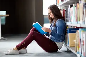 A woman sits on the floor in a library reading a book
