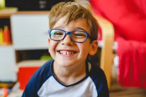 A boy wearing glasses is smiling and looking at the camera. He is in a room with a wooden chair and a shelf behind him.