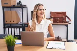 woman wearing glasses sitting at a desk with a laptop in an office