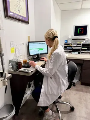 A woman in a white lab coat is working at a computer in a medical office.