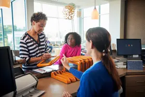 Three women are in an office with a monitor, printer, and glass windows. One woman is explaining something to the other two women.