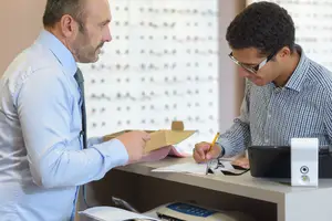 A man wearing glasses is writing on a piece of paper at a counter while another man in a blue shirt is looking at him.