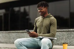 A man wearing a green hoodie, glasses, and jeans is sitting on the steps and smiling while looking at his cellphone.