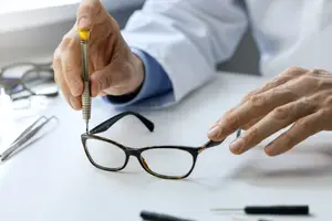 A person is fixing a pair of glasses using a screwdriver and other tools on a white table.