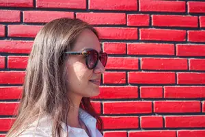 Close-up of a woman with long hair wearing sunglasses, smiling and looking sideways against a red brick wall.