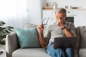 An elderly man sitting on a couch with his hands on his face while holding his glasses.