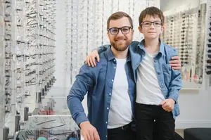 A man and a boy are standing next to each other in a store with eyeglasses on display behind them.