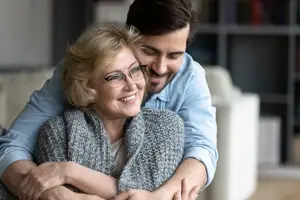 An elderly woman and a young man are smiling and hugging each other in a living room