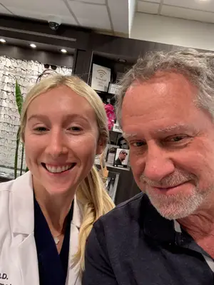 A man and woman are smiling at each other in a room with a cabinet full of eyeglasses and a surveillance camera on the ceiling.