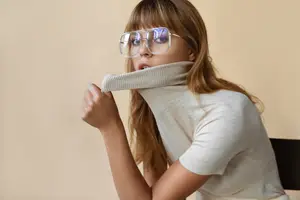 A woman is adjusting her turtleneck shirt and looking at something with her mouth open. She is wearing glasses and sitting on a chair with her hands on her neck. She is probably posing for a photo with a blurred background.