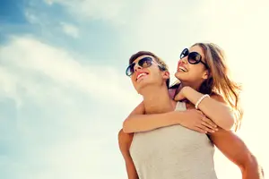 A couple enjoying a day at the beach, the woman is giving a piggyback ride to the man who is smiling and looking at the camera