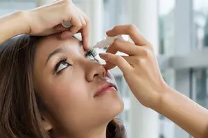 A woman is applying eye drops in a room with windows and a blurred view of the outside