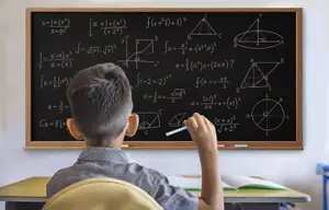 A young boy is sitting at a desk in front of a blackboard, holding a pen and pointing at the board with his left hand while seemingly solving a math problem. He has a book and a pen on the desk in front of him.