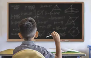 A boy sitting in a classroom raising his hand to answer a question on a blackboard with mathematical equations and geometric shapes