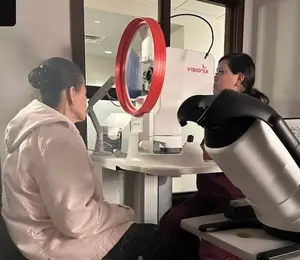 Two women are in an eye care clinic, one is sitting on a chair and the other is standing in front of an eye exam machine.