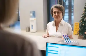 An older woman in glasses sitting at a reception desk with a monitor, smiling while looking at someone in front of her.