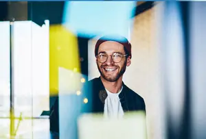 A smiling man wearing a beanie and glasses standing in an office room with glass windows and a blurry background