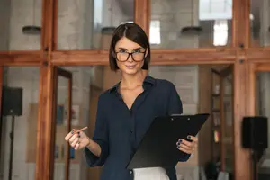 A woman wearing glasses and a blue shirt holding a clipboard and pen is smiling while standing in an indoor area