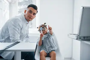 A young girl with glasses on her face is sitting on a chair in an optometrist's office, and a man in a white coat is sitting in front of her and looking at her glasses.