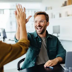 A smiling man in glasses in an office room gives a high five to a woman in a yellow shirt.
