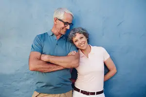 An elderly couple smiling and posing for a picture against a blue wall.