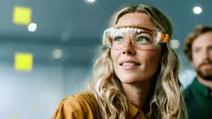 A woman with blonde hair wearing safety goggles is standing in a room with a man in a green shirt behind her.