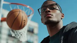 A man wearing glasses is looking up at a basketball that is in the air, possibly about to be shot into the basketball hoop in the background.
