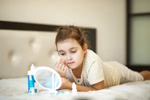 A young girl lying on a bed is applying something to her face with her right hand and holding a mirror in her left hand.