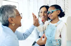 A girl is wearing goggles and holding a stick while a woman in glasses and a woman are smiling and looking at her.
