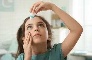 A girl wearing a green dress is applying eye drops to her eye while looking up