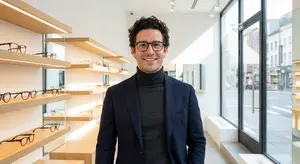 A man wearing glasses and a suit stands in front of a shelf of glasses in a store.