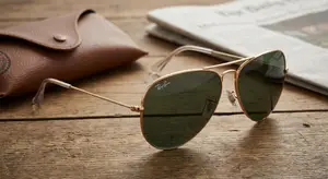 Ray-Ban Aviator sunglasses with brown lenses on a wooden table next to a brown leather case and a newspaper
