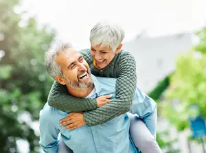 An elderly man and woman are outside on a sunny day, the woman is sitting on the man's shoulders and smiling, the man is looking at her and smiling back