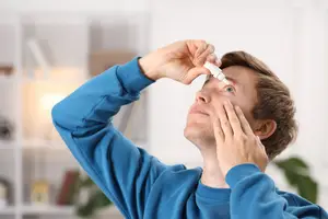 A man wearing a blue long-sleeve shirt is holding a white bottle of eye drops in his hand and seems to be applying it to his eye.