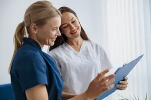 Two women in blue uniforms discussing something with a clipboard in hand.