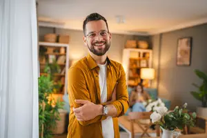 A man standing in a living room with his arms crossed and a woman sitting on a couch behind him.