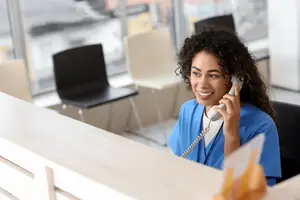 A woman in a blue scrubs is sitting at a desk, holding a phone to her ear, and smiling at the camera.