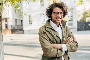 A smiling man in a green jacket and glasses stands with his arms crossed on a city street.