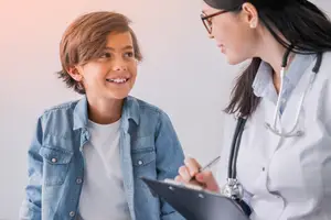 A smiling young boy is looking at the doctor while holding a clipboard and a pen