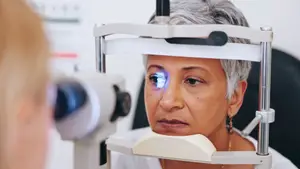 An elderly woman is undergoing an eye examination, looking into an eye examination machine while being examined by another person, both are wearing eyeglasses.