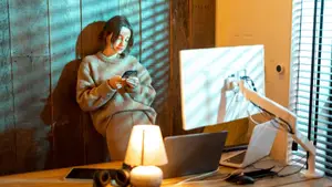 A woman sitting in front of a desk with a computer monitor, laptop, and cell phone.