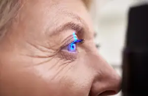 An elderly woman with blue light illuminating her eye is being examined by a doctor using an ophthalmoscope in a hospital setting