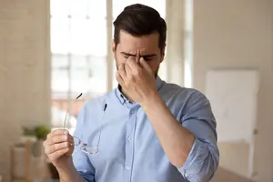 A man wearing a blue shirt is holding a pair of eyeglasses and touching his forehead with his left hand