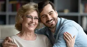 A smiling elderly woman wearing glasses and a man in a blue shirt are hugging and smiling on a couch