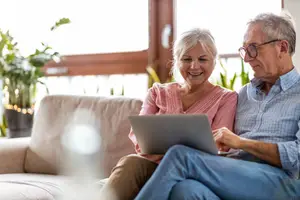 Senior couple sitting on a couch using a laptop and smiling at the camera