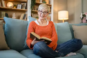 An elderly woman sitting on a couch, wearing glasses, reading a book, with a shelf and lamp in the background.