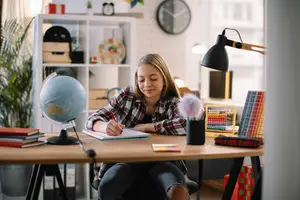 A girl is sitting on a chair and seems to be studying on the table. A lamp is placed on the desk beside her. A globe, books, a pen stand, and a pencil box are on the desk. A clock, a storage box, a hat, and a potted plant are on the shelf. A frame is mounted on the wall. A glass window is beside the girl.