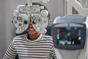 A young girl wearing a striped shirt is sitting in front of an ophthalmoscope machine and smiling.