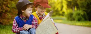 Two young boys are sitting on the grass, one of them reading a book and the other smiling and looking at him, with trees and plants in the background.